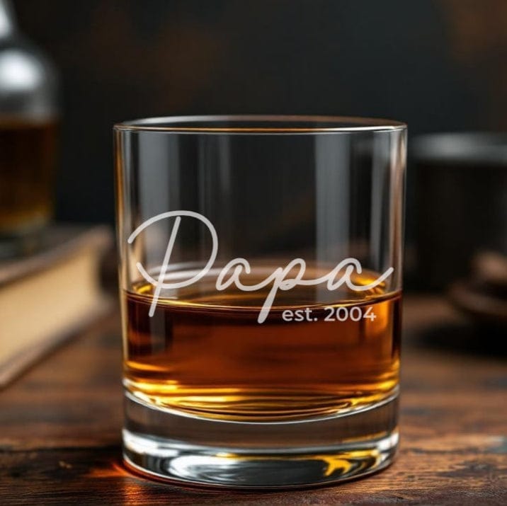 Glass of whiskey with 'Papa est. 2004' engraving on a wooden table.