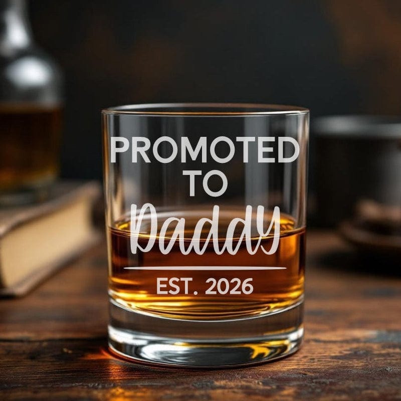 Glass of whiskey with 'Promoted to Daddy Est. 2026' etched text on a wooden surface.