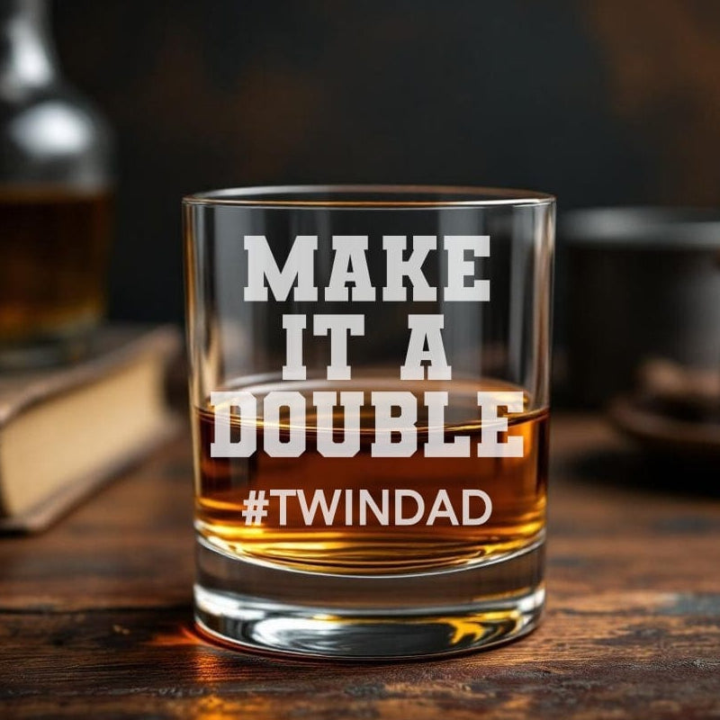 Glass of whiskey with text 'MAKE IT A DOUBLE #TWINDAD' on a wooden surface.