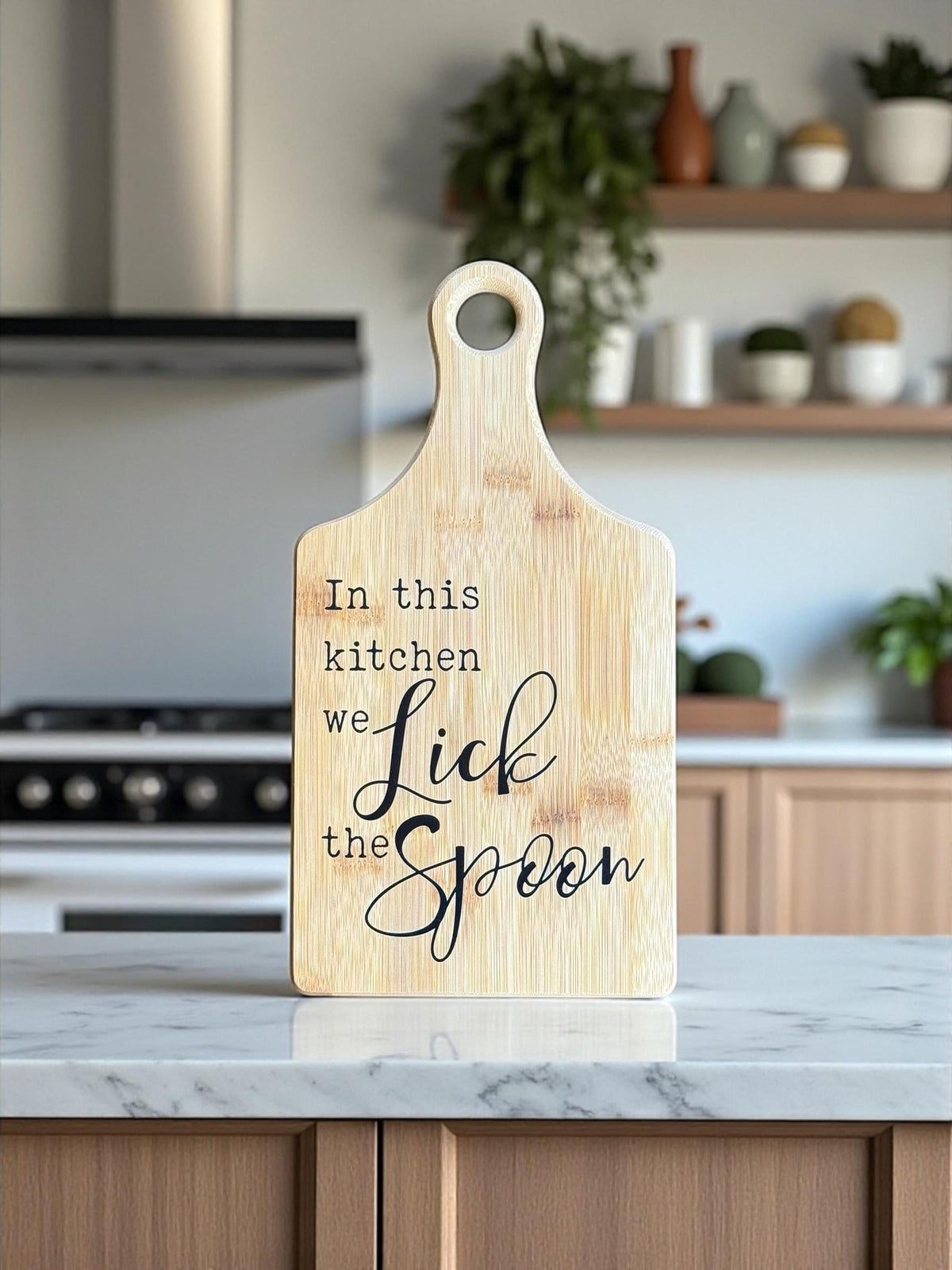Decorative bamboo paddle board with baking-themed lettering and the words “In This Kitchen We Lick the Spoon”.