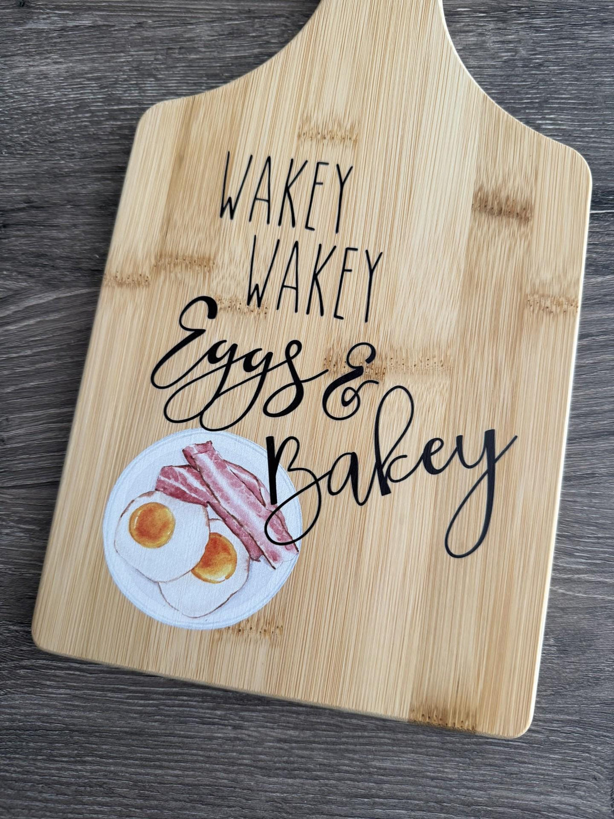 Decorative bamboo paddle board with breakfast-themed lettering and the words “Wakey Wakey Eggs and Bakey”.