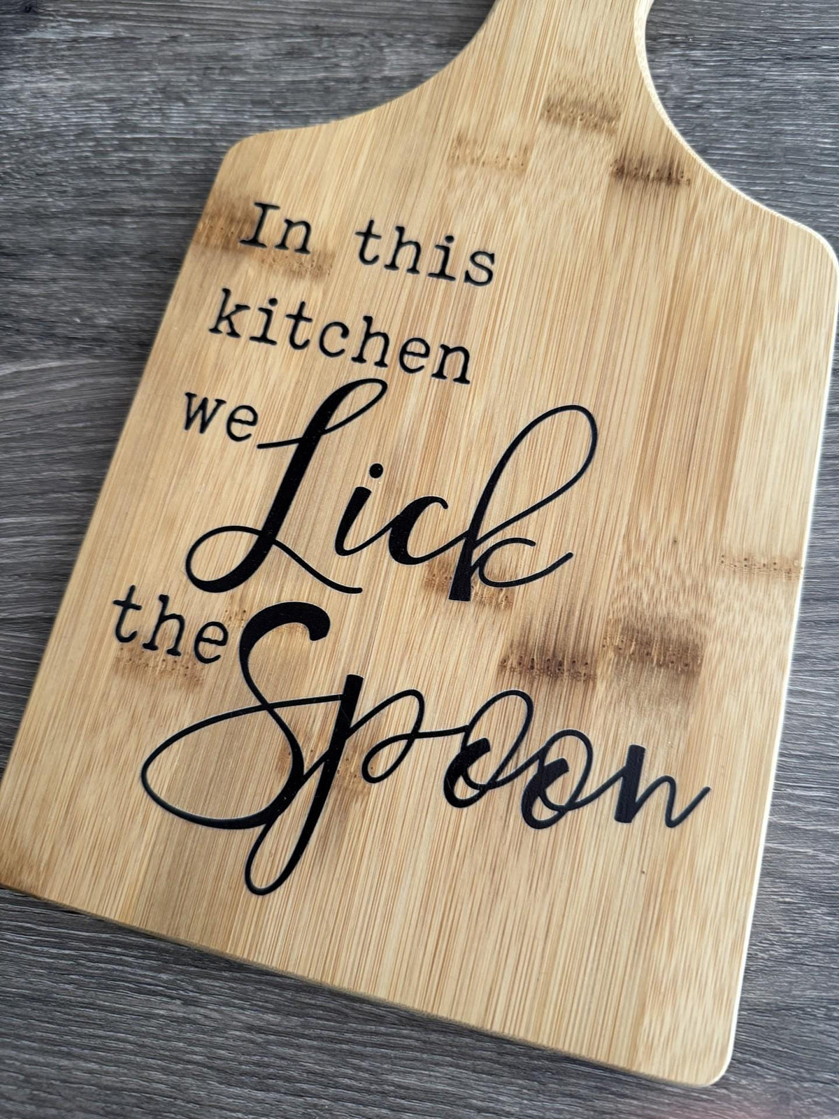 Decorative bamboo paddle board with baking-themed lettering and the words “In This Kitchen We Lick the Spoon”.