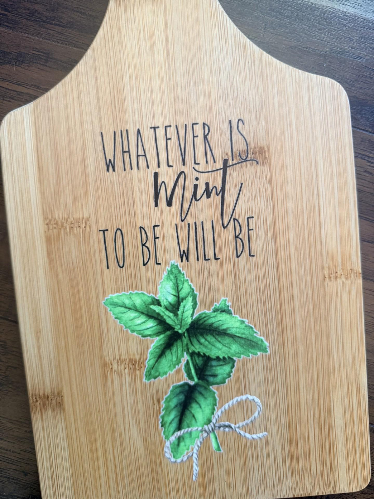 Decorative bamboo paddle board with mint herb sprig and the words “Whatever Is Mint to Be Will Be”.