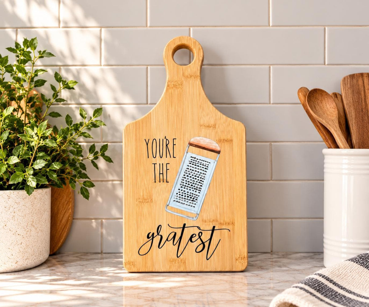 Decorative bamboo paddle board with cheese grater illustration and the words “You're the Gratest”.