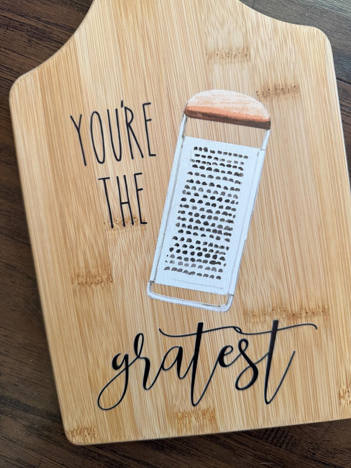 Decorative bamboo paddle board with cheese grater illustration and the words “You're the Gratest”.