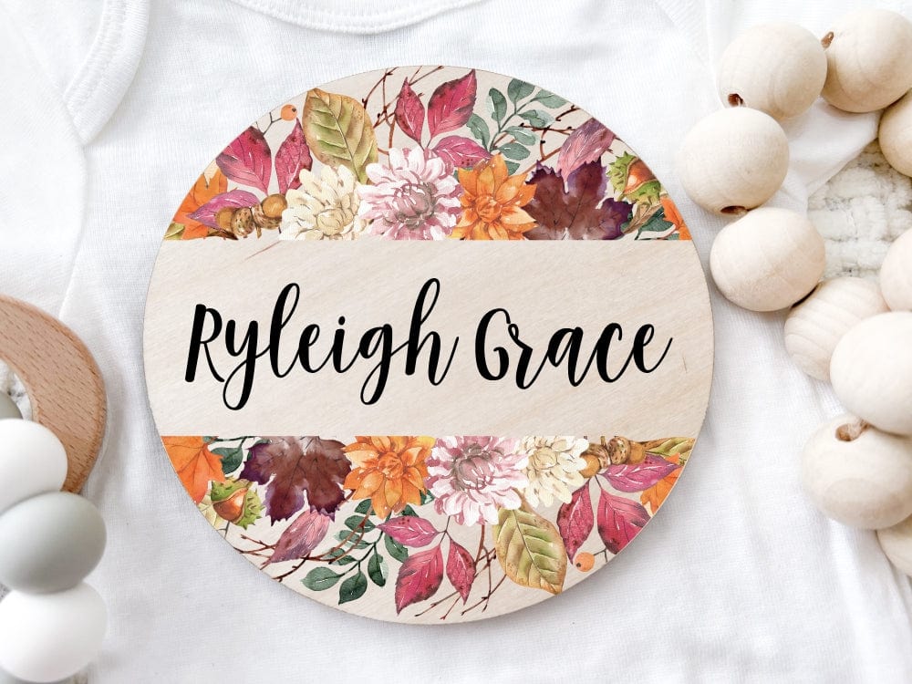round wood baby name sign uv printed with fall like florals and leaves in bold colors at the top and bottom personalized with baby first and middle name in center