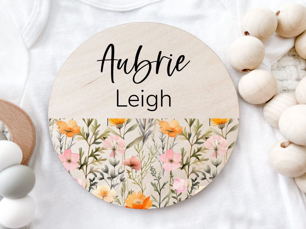 round wood baby name announcement sign printed with baby name and bottom half of sign has a floral pattern with yellow/orange and pink flowers and greenery
